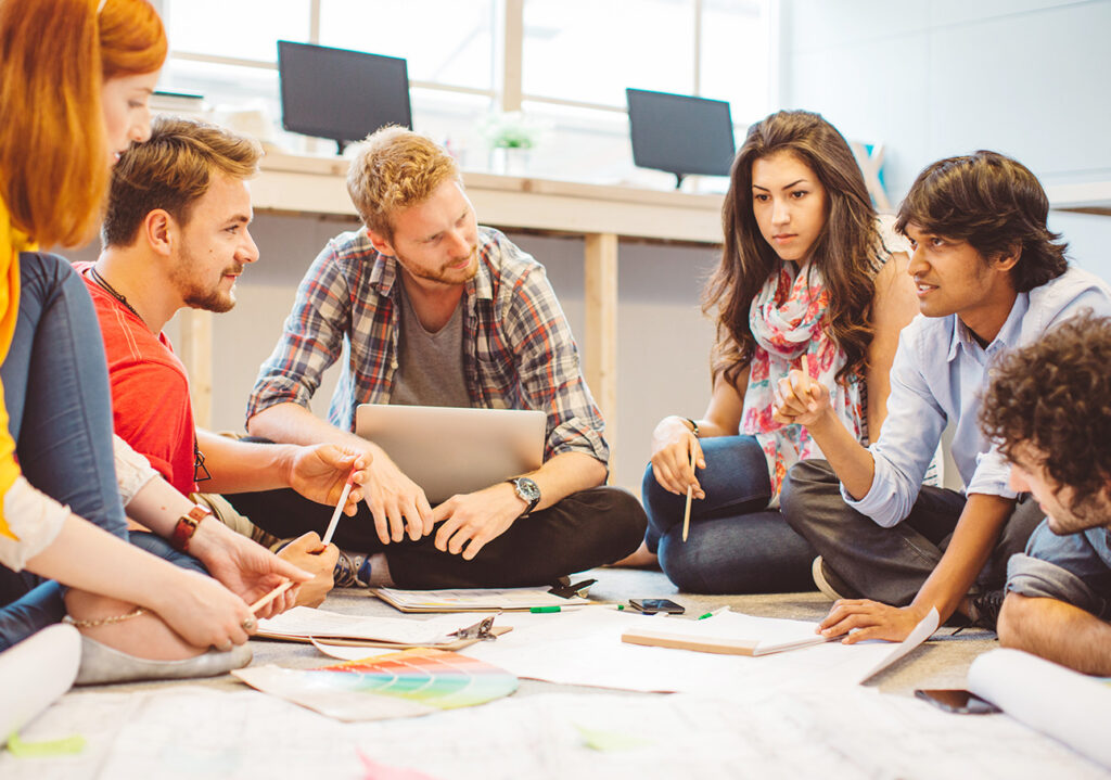 Gruppe von jungen Studenten und Studentinnen sitzen im Kreis in einem Raum, vor Ihnen liegen Zeichnungen und Mitschriften. Sie planen ein Projekt und sehen konzentriert aus